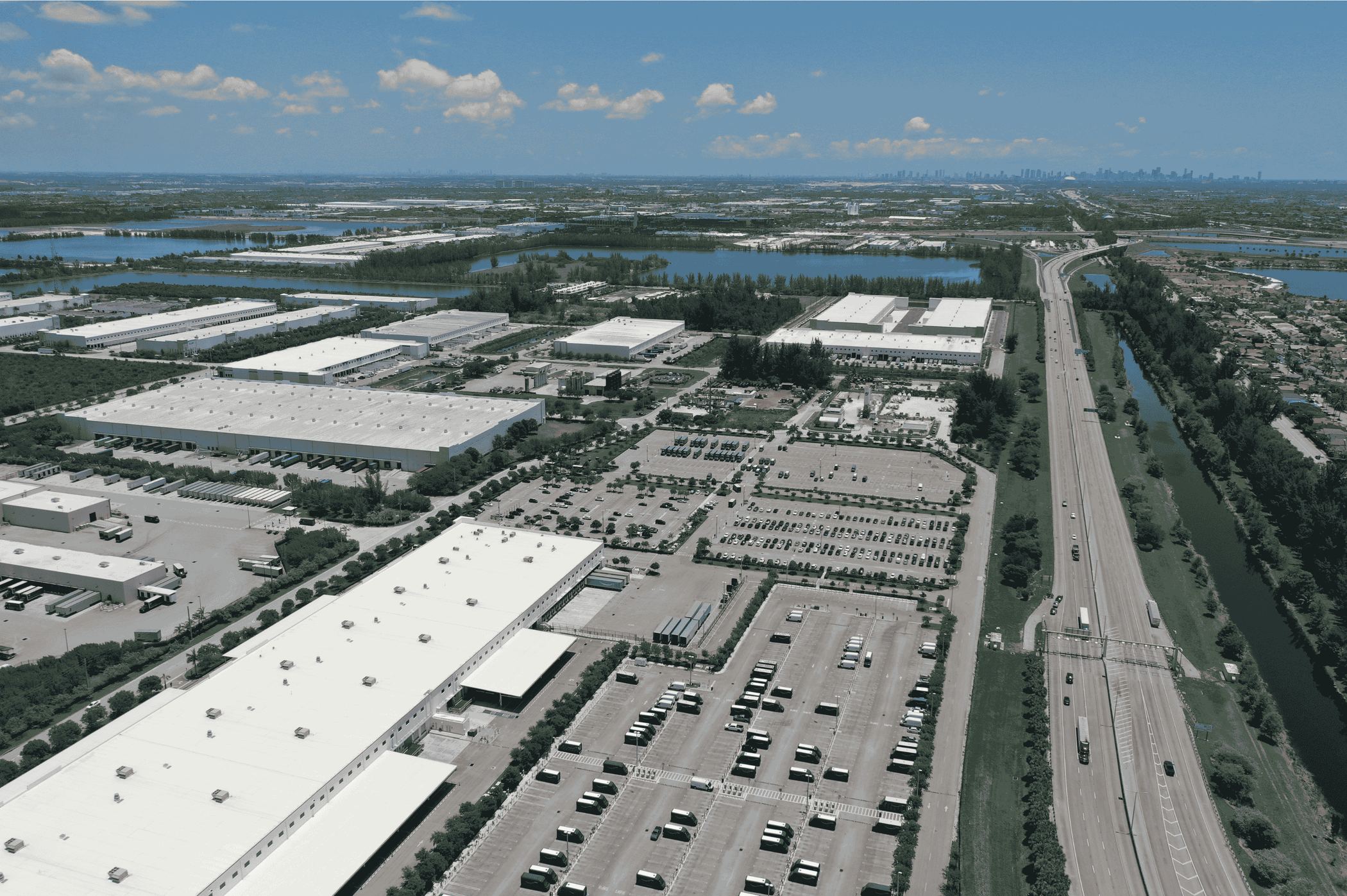 Aerial view of a Miami-Dade industrial park with warehouses, truck and van yards, and a highway, with the downtown Miami skyline on the horizon