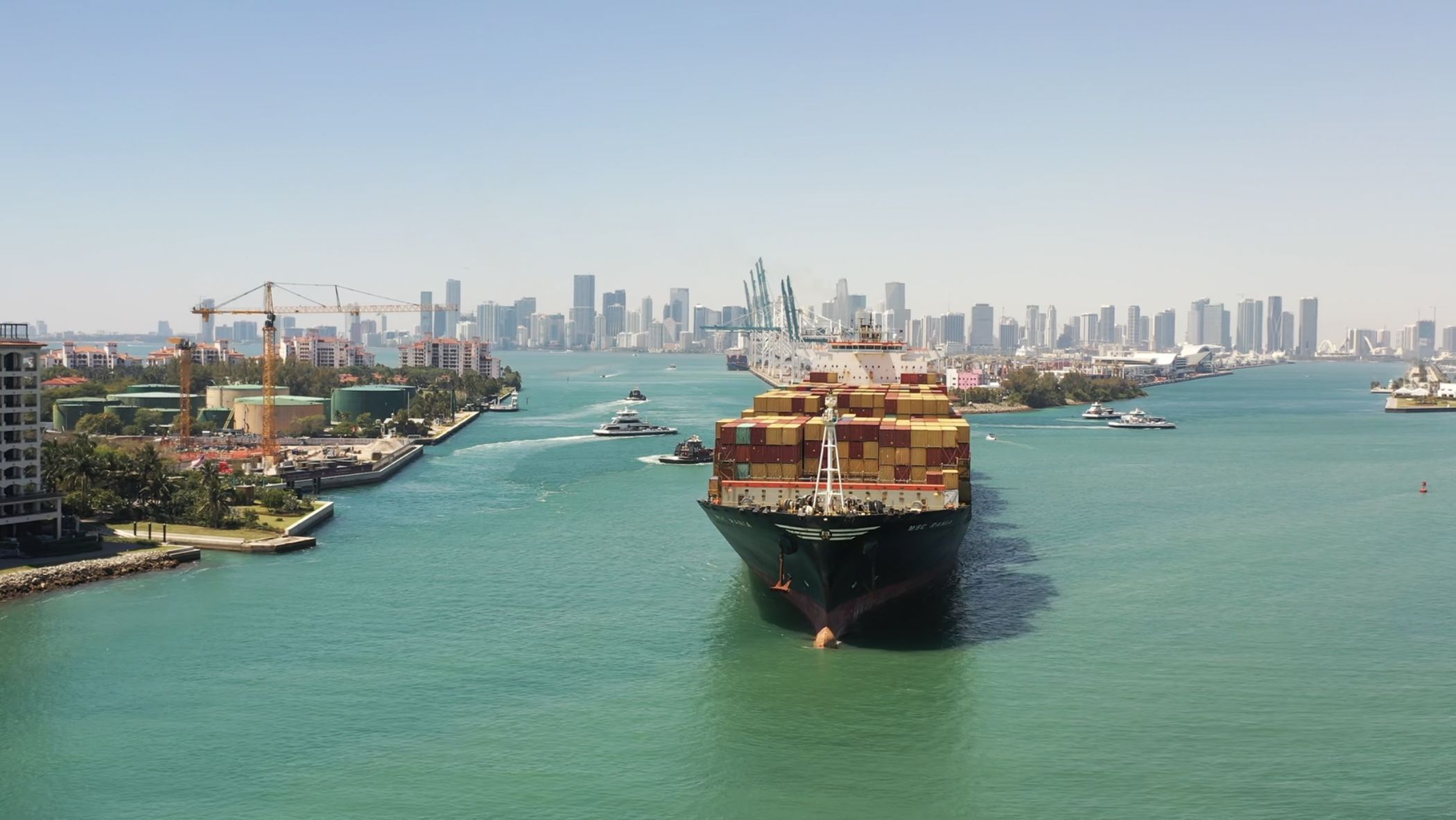 Loaded container ship entering PortMiami through Government Cut, with the port's cranes and the downtown Miami skyline behind it